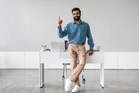 Photo for Handsome indian businessman sitting leaning on desk at workplace and pointing finger up at free place, smiling at camera. Man in formalwear showing copy space for your ad or text - Royalty Free Image