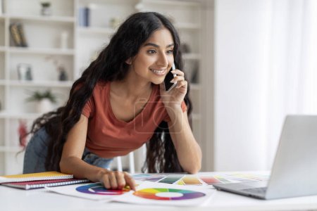 Photo for Freelance Designer Young Indian Woman Balancing Tech and Talk. Energized and spirited, lady designer balancing between using laptop and phone conversation with client, home workspace interior - Royalty Free Image