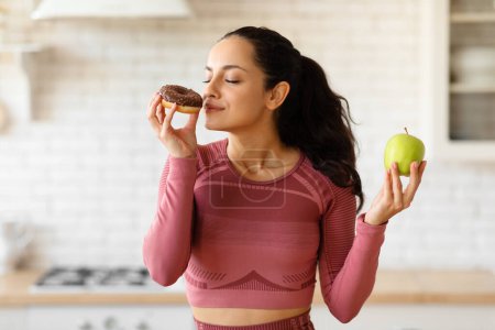Photo for Fitness woman choosing a sugary donut instead of healthy apple in modern kitchen, sniffing tasty doughnut while standing in fitwear. Food choice and healthy eating routine concept - Royalty Free Image