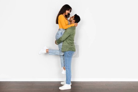 Photo for Young indian man holds his wife aloft in his arms, both sharing genuine, joyful smiles, creating uplifting image of love, support, and playful togetherness at home - Royalty Free Image