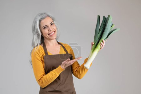 Photo for Smiling pretty calm senior woman farmer in apron point hand on onion leek, enjoy eco green harvest, isolated on gray background. Healthy food recommendation, agro business, farmer work, ad, offer - Royalty Free Image
