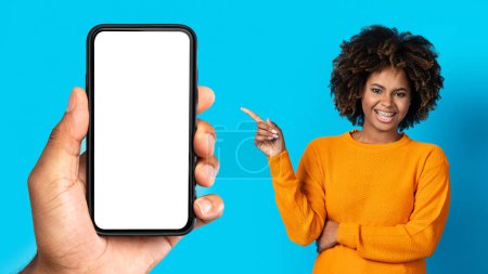 Photo for Smiling curly millennial african american lady with teeth braces pointing finger at hand with phone with empty screen, isolated on blue studio background, panorama. App recommendation - Royalty Free Image