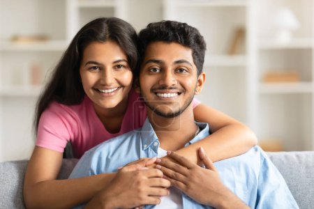 Foto de Joven pareja india comparte abrazo apretado, sintiendo felicidad, sonriendo a la cámara, unidos en el amor, sentado en el sofá en el interior de casa, primer plano - Imagen libre de derechos