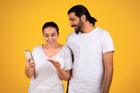 Photo for Smiling arab adult lady pointing hand at smartphone, sharing exciting content, isolated on yellow studio background. Expressions filled with curiosity and amusement with device - Royalty Free Image
