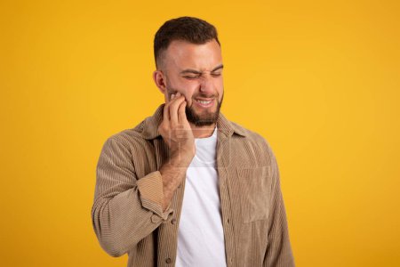 Photo for Despaired millennial european guy with beard put hand to cheek, suffering from toothache, isolated on orange studio background. Caries, emotions, medicine, dentistry problems - Royalty Free Image