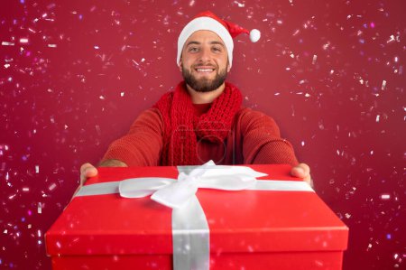 Photo for Smiling millennial caucasian man with beard in Santa hat give box present with snow, confetti, isolated on burgundy studio background, close up. Christmas, New Year holidays, ad and offer, party - Royalty Free Image