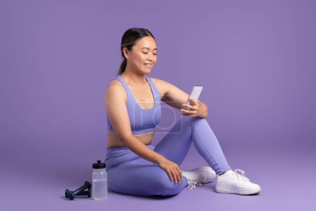 Téléchargez les photos : Femme asiatique en vêtements de sport s'engage avec son téléphone portable, assis et se reposant post-entraînement sur le sol, sur fond de studio violet, illustrant une pause tranquille - en image libre de droit