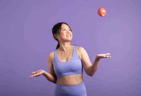 Photo for Active asian woman in sportswear tossing red apple, creating a dynamic and playful scene in purple studio background. Sports, fitness, body care and vitamins food - Royalty Free Image