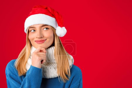 Téléchargez les photos : Femme rêveuse dans le chapeau de Père Noël regardant vers le haut à l'espace de copie et touchant menton sur fond rouge, penser aux cadeaux célébration de Noël, bannière - en image libre de droit