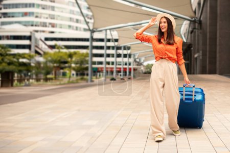 Foto de Joven dama sonriente con maleta de pie al aire libre en la moderna terminal del aeropuerto. Mujer viajera posando con su equipaje esperando el embarque de vuelo, lista para las vacaciones de verano - Imagen libre de derechos