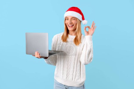 Foto de Compras de Navidad en línea. Feliz dama europea en Santa sombrero sosteniendo ordenador portátil y mostrando signo ok, aprobando algo y sonriendo en fondo de estudio azul - Imagen libre de derechos