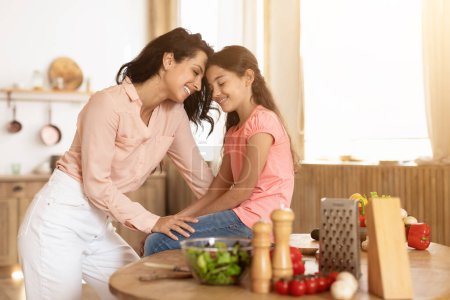 Foto de Mamá y su hijita comparten un momento sincero tocando la frente mientras preparan una ensalada saludable, apreciando el amor familiar y el vínculo durante la preparación de la cena en la cocina en casa - Imagen libre de derechos