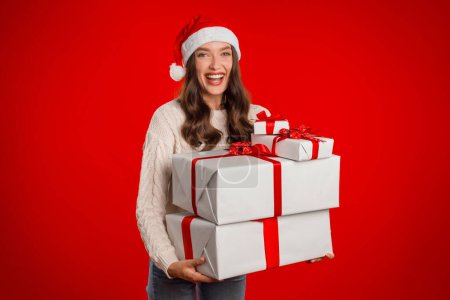 Téléchargez les photos : Joyeux jeune femme dans le chapeau de Père Noël tenant deux boîtes cadeaux de Noël sur fond de studio rouge, saluant exprimant la positivité et joyeux sentiments de vacances. Livraison et célébration de cadeaux de Noël - en image libre de droit