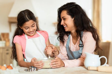 Foto de Alegre mamá y su hijita disfrutando de la preparación de masa de galletas, pastelería para hornear en la cocina el fin de semana. Mamá y niño chica vinculación durante la cocina de ocio, amasar y mezclar ingredientes - Imagen libre de derechos