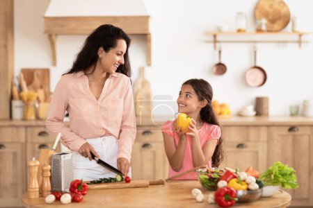Foto de Sonriente madre e hija preparando ensalada para la cena en la cocina moderna en casa, cortando verduras orgánicas frescas. Tiempo familiar de calidad con una preparación nutritiva de alimentos - Imagen libre de derechos
