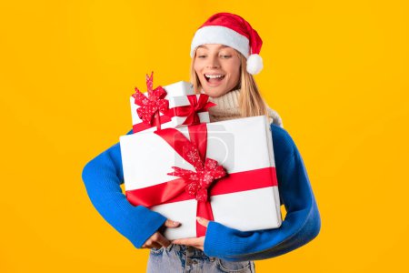 Téléchargez les photos : Femme européenne excitée en pull et chapeau Santa tenant pile de cadeaux, dame avec tas de cadeaux célébrant les vacances d'hiver sur fond jaune studio - en image libre de droit