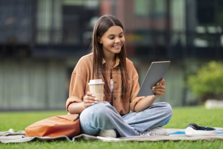 Photo for Relaxed cheerful young woman student chilling on green lawn at university campus, have break, drink takeaway coffee, using digital tablet, enjoy video call with friends. Communication and gadgets - Royalty Free Image
