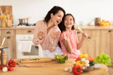 Foto de Alegre mamá e hija niño usando cucharas como micrófonos, cantando mientras hacen ensalada fresca juntos, de pie cerca de una mesa con verduras orgánicas en la cocina moderna, disfrutando de la diversión de la cocina familiar - Imagen libre de derechos