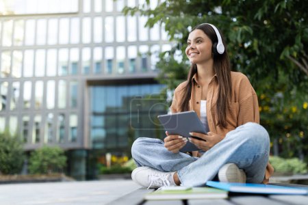 Photo for Cheerful pretty young woman student listening to music outdoors, using wireless headphones and digital tablet, enjoying favorite podcast while have break at college, looking at copy space - Royalty Free Image