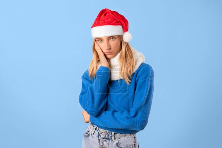 Foto de Mujer joven infeliz en Santa sombrero tocando mejilla y mirando a la cámara con expresión entristecida, contrastando vibrante atuendo de vacaciones contra el fondo del estudio azul - Imagen libre de derechos