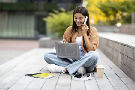 A joyous woman student at a barren university spot engages in a phone conversation and peruses online study materials via her laptop, fully immersed in remote education, copy space
