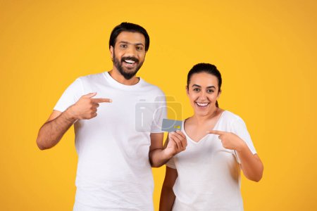 Photo for Cheerful millennial arab couple point finger at credit card, recommends sale, profit and savings, isolated on yellow studio background. Online shopping platform, credit advice - Royalty Free Image