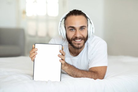 Téléchargez les photos : Jeune homme souriant utilisant une tablette numérique avec écran blanc vide tout en étant couché sur le lit le matin, en utilisant l'application mobile sur pad et casque sans fil, espace de copie, intérieur de la maison, maquette - en image libre de droit