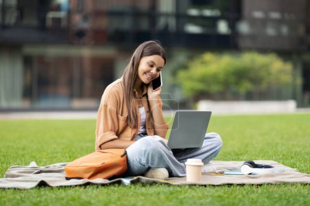 Photo for Happy pretty young woman student, amidst a silent, empty university setting, animatedly talks on her phone, while her other hand navigates through an online educational portal on her laptop - Royalty Free Image