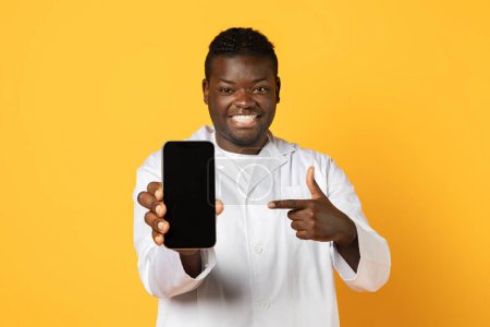 Foto de Aplicación médica. Sonriente afroamericano médico en uniforme que demuestra el teléfono inteligente en blanco en la cámara, médico que muestra la pantalla vacía del teléfono celular en la mano sobre el fondo amarillo, burla - Imagen libre de derechos
