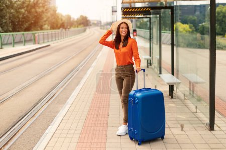 Photo for Tourist woman in summer hat with luggage waiting for the tram transport at public stop outdoor, standing with big blue suitcase travelling through urban city via modern streetcar. Empty space - Royalty Free Image