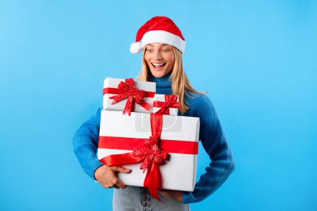 Téléchargez les photos : Femme excitée dans le chapeau du Père Noël tenant de nombreuses boîtes cadeaux, debout sur fond bleu studio. Vente de vacances d'hiver et offre de livraison de Noël - en image libre de droit