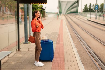 Téléchargez les photos : Jeune femme décontractée parlant au téléphone et attendant un tram à l'arrêt, debout avec une grande valise de voyage bleue, vue de côté. Espace gratuit pour la publicité des transports publics - en image libre de droit