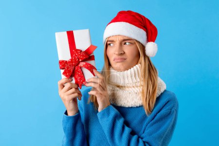 Téléchargez les photos : Femme européenne déçue tenant une boîte cadeau, regardant la boîte être malheureuse avec le cadeau, dame portant le chapeau de Père Noël sur fond bleu. Concept de Noël - en image libre de droit