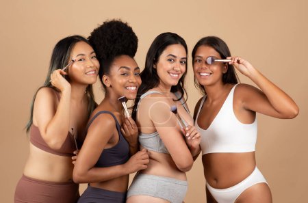 Foto de Cuatro mujeres multirraciales viga con sonrisas alegres, cada uno con pinceles de maquillaje, presentando colectivamente viva instantánea de la unidad y la belleza variada en el estudio beige - Imagen libre de derechos