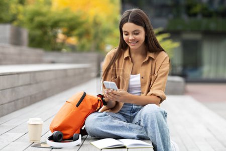 Photo for Joyful pretty young woman student browsing with a beaming smile on her smartphone, sitting relaxed at the serene, green university campus, embodying a peaceful academic break, nice mobile app - Royalty Free Image
