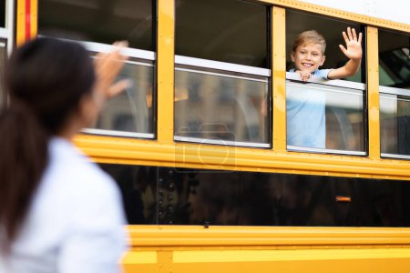 Foto de Niño sentado dentro del autobús escolar amarillo y saludando de la mano a la madre, niño feliz emocionado asomándose por la ventana del vehículo y diciendo adiós a mamá, listo para el viaje a la escuela, enfoque selectivo - Imagen libre de derechos