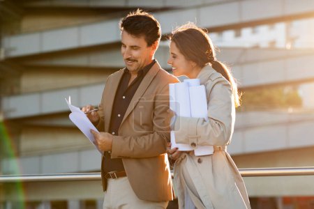 Photo for A man and woman, mature business partners reading construction drawings while standing on a vibrant city street, reflecting focused collaboration and proactive discussions, sun flare - Royalty Free Image
