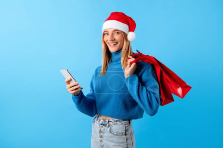 Foto de Joven mujer europea en Santa sombrero sosteniendo bolsas de compras y el uso de teléfonos inteligentes, compras en Internet, mientras que las ventas de Navidad, de pie sobre fondo de estudio azul - Imagen libre de derechos