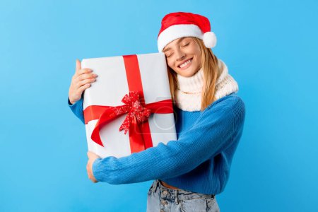 Photo for Excited caucasian woman in Santa hat holding and hugging big wrapped gift box over blue studio background. Happy lady celebrating New Year or Christmas - Royalty Free Image