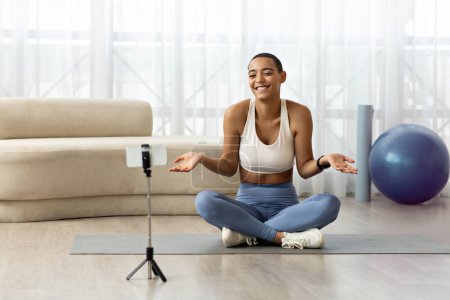 Photo for Positive confused millennial latin lady blogger in sportswear sit on floor, shooting video fitness blog on phone in living room interior. Weight loss and sports at home, fitness - Royalty Free Image