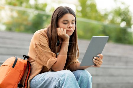 Téléchargez les photos : Jeune étudiante énervée utilisant une tablette numérique à l'extérieur, assise sur le campus universitaire ou dans un parc public seule, regardant l'écran de la tablette, se sentant ennuyée ou seule, éprouvant des difficultés à faire ses devoirs - en image libre de droit