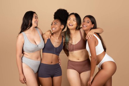 Foto de Diversas mujeres muestran su belleza natural en ropa interior discreta, compartiendo un momento de alegría y unidad, contra el fondo beige del estudio - Imagen libre de derechos
