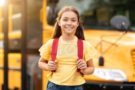 Foto de Feliz sonrisa niña preadolescente de pie cerca de autobús escolar amarillo al aire libre, alegre lindo niño femenino con mochila posando fuera, listo para las clases, hermosa colegiala mirando a la cámara, disfrutando del estudio - Imagen libre de derechos