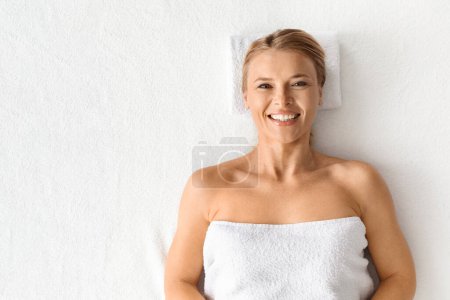 Photo for Beautiful middle aged woman enjoys her time at luxury spa salon, attractive mature lady wrapped in towel lying comfortably on a table and smiling at camera, top view with copy space - Royalty Free Image