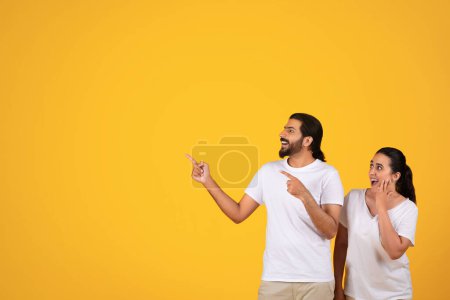 Photo for Glad shocked millennial arab couple pointing fingers at empty space, for text or ad, isolated on yellow studio background. Attention towards vacant area, recommendation sale, surprise - Royalty Free Image