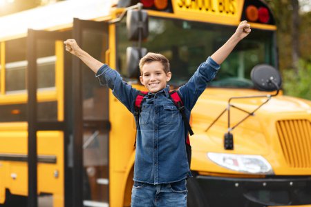 Foto de Feliz niño preadolescente excitado levantando brazos mientras está parado cerca del autobús escolar amarillo, alegre alumno masculino con mochila lista para el estudio, niño positivo disfrutando de un nuevo día en la escuela, espacio de copia - Imagen libre de derechos