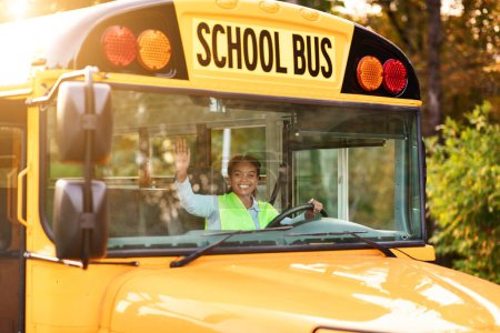 Photo for Portrait of joyful young black female driver steering yellow school bus, happy african american woman in uniform sitting on drivers seat and waving hand at camera, driving along the street - Royalty Free Image