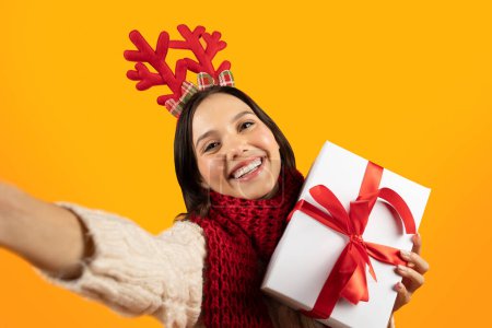 Photo for Cheerful Young Lady With Festive Deer Horns And Wrapped Xmas Present Taking Selfie, Enjoying Winter Holidays Posing On Yellow Background, Studio Shot. Christmas Fun And Gifts - Royalty Free Image