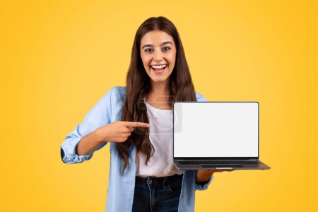 Foto de Sorprendido contento europeo adolescente estudiante dama señalar el dedo en el ordenador portátil, aislado en el fondo amarillo, estudio. Estudie recomendaciones remotas, educación y lecciones en línea, cursos de atención - Imagen libre de derechos