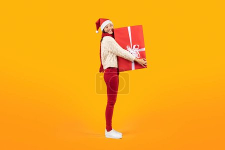 Photo for Full length of young woman wearing Santa hat and holding Christmas gift box over yellow studio backdrop, smiling to camera. Joy and kindness of holiday celebrations, Xmas presents season - Royalty Free Image